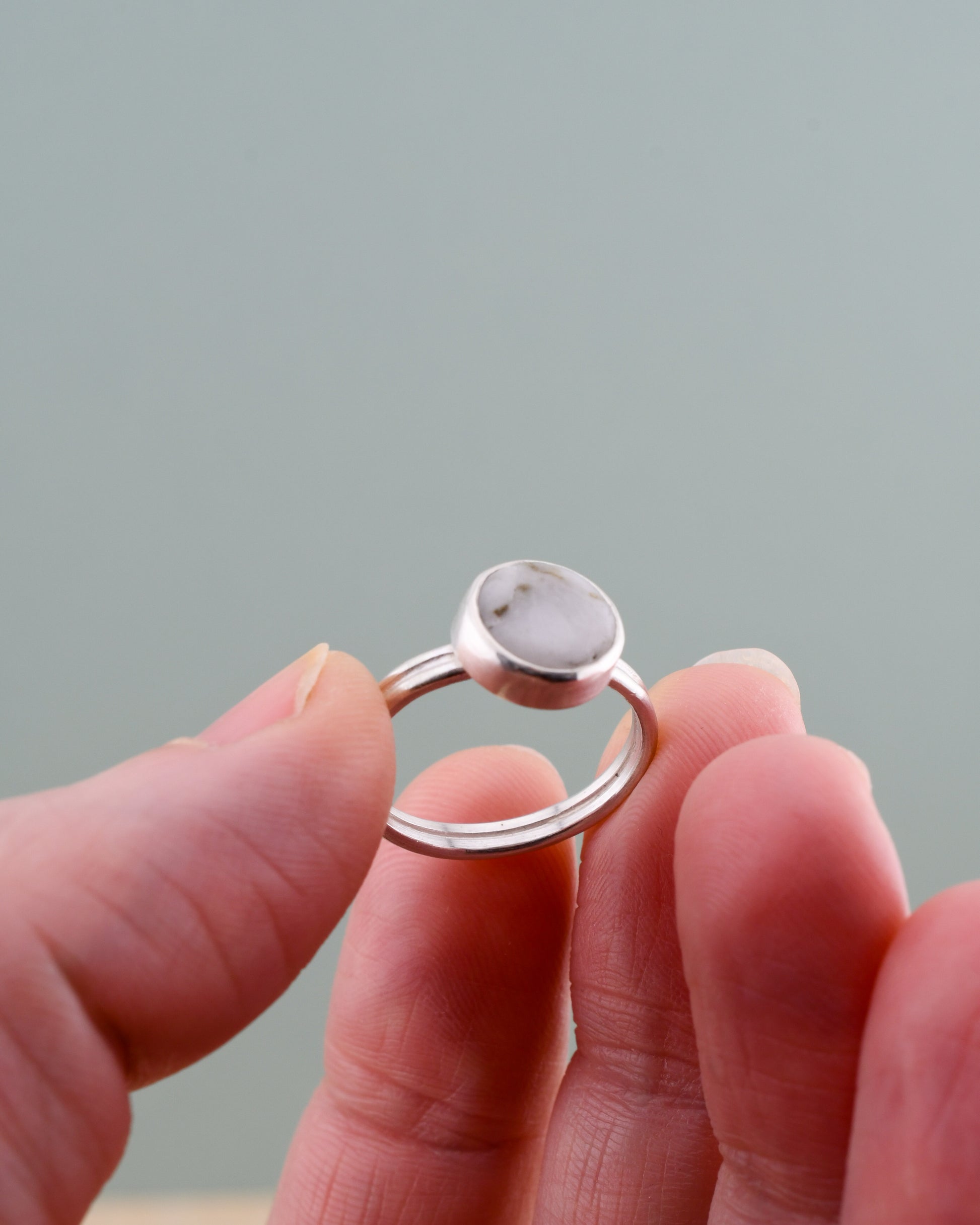 Silver ring with white stone held between fingers against a neutral background