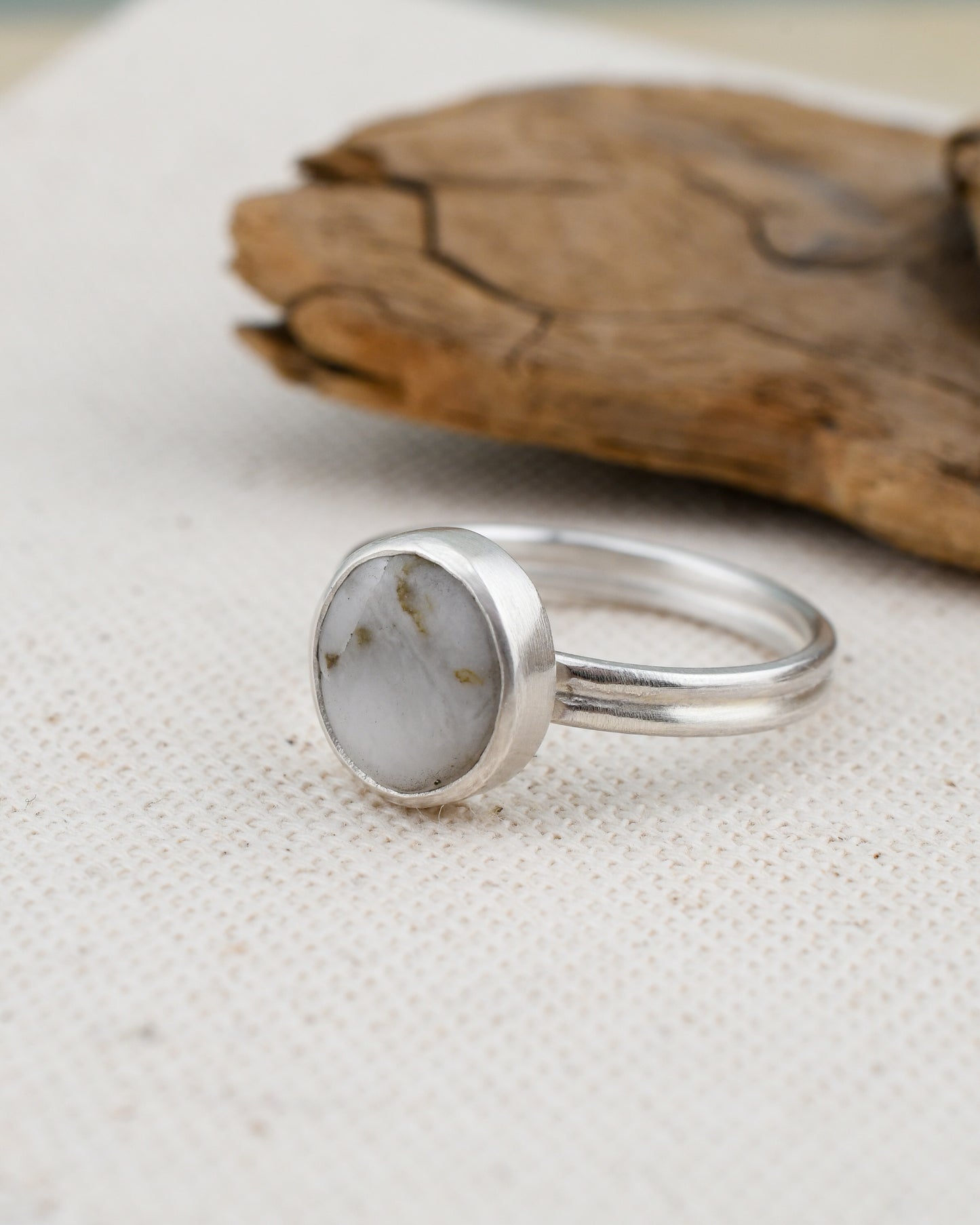Silver ring with a round white stone on a textured beige surface with a wooden object in the background