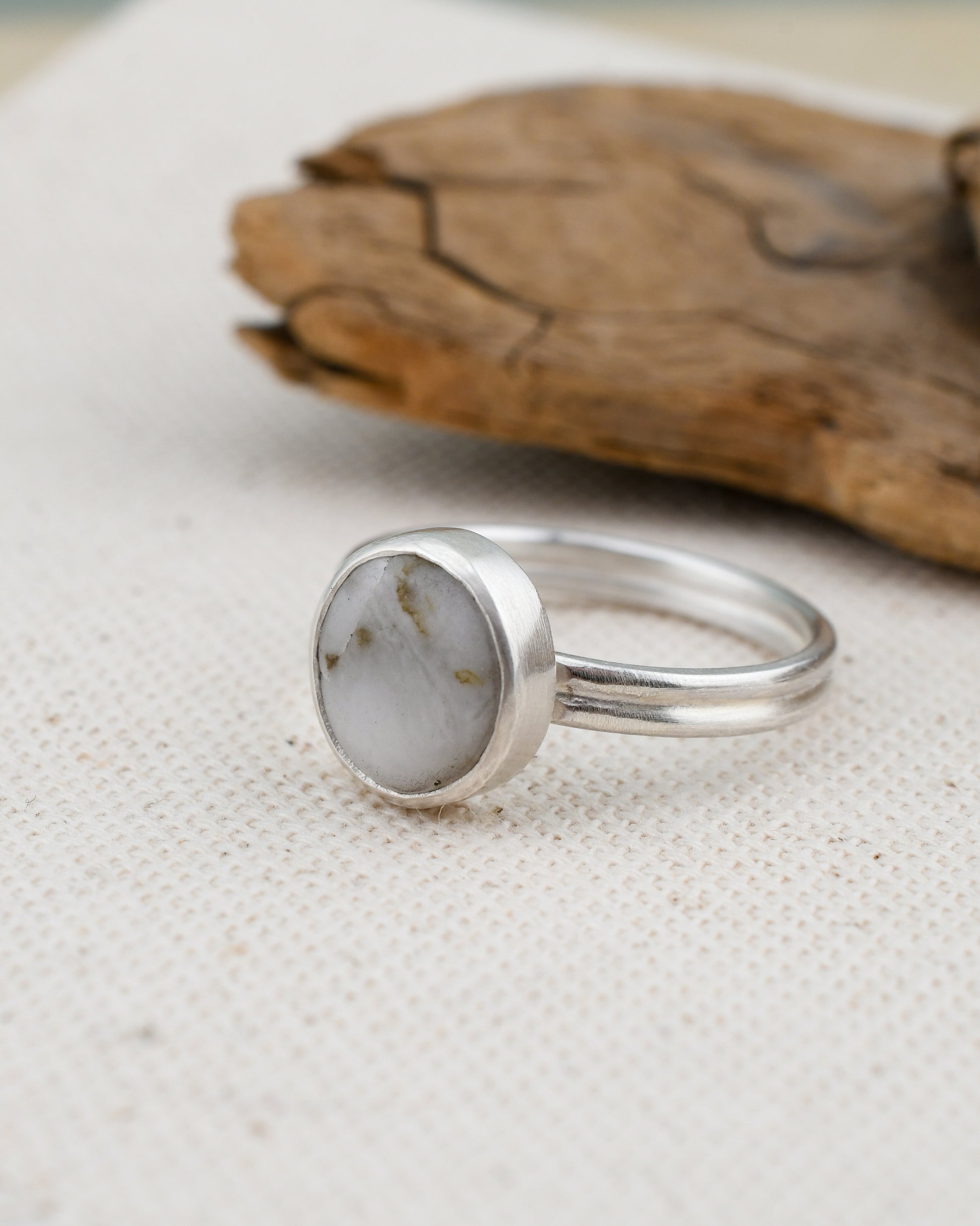 Silver ring with a round white stone on a textured beige surface with a wooden object in the background