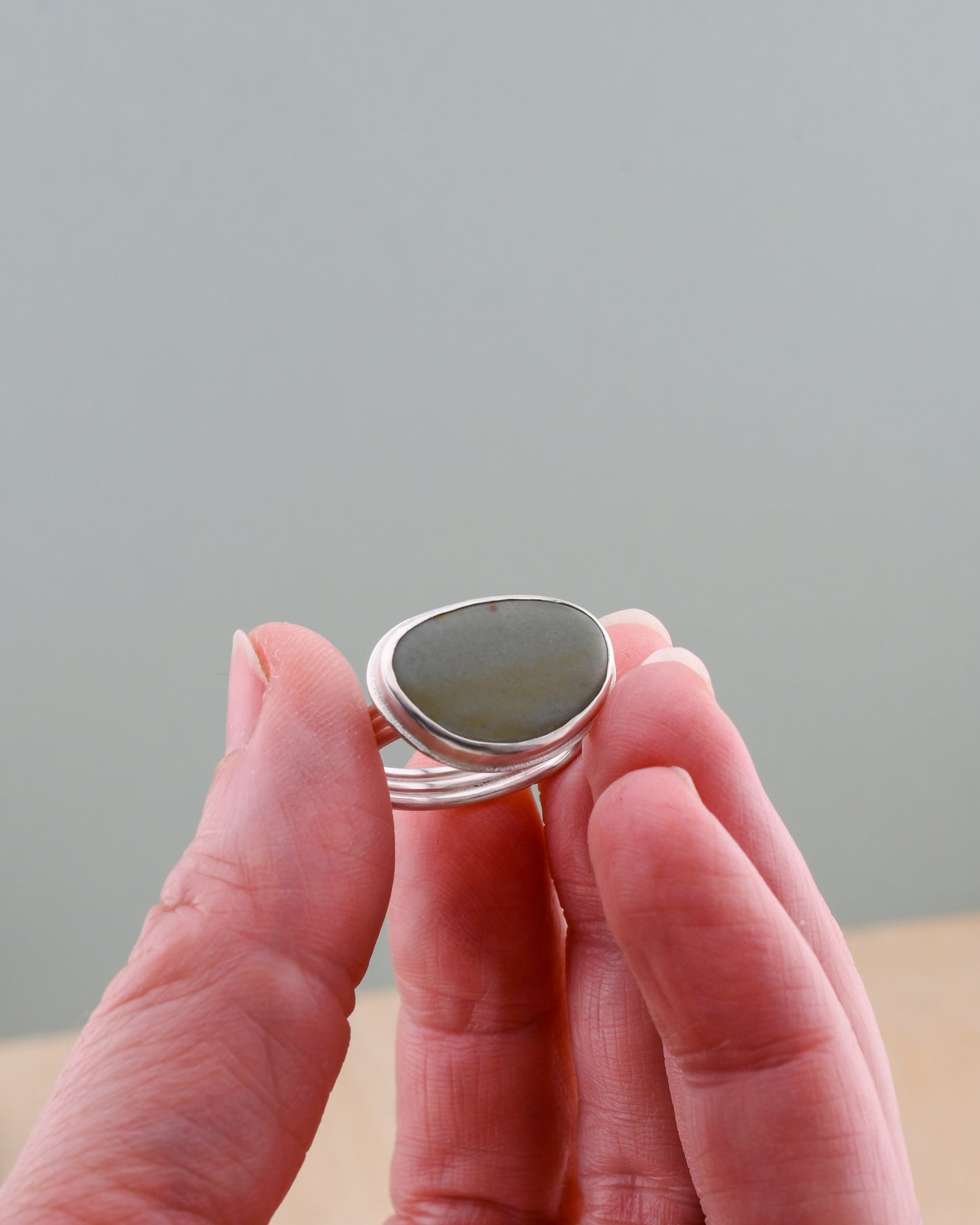 Hand holding a silver ring with a beach stone against a neutral background