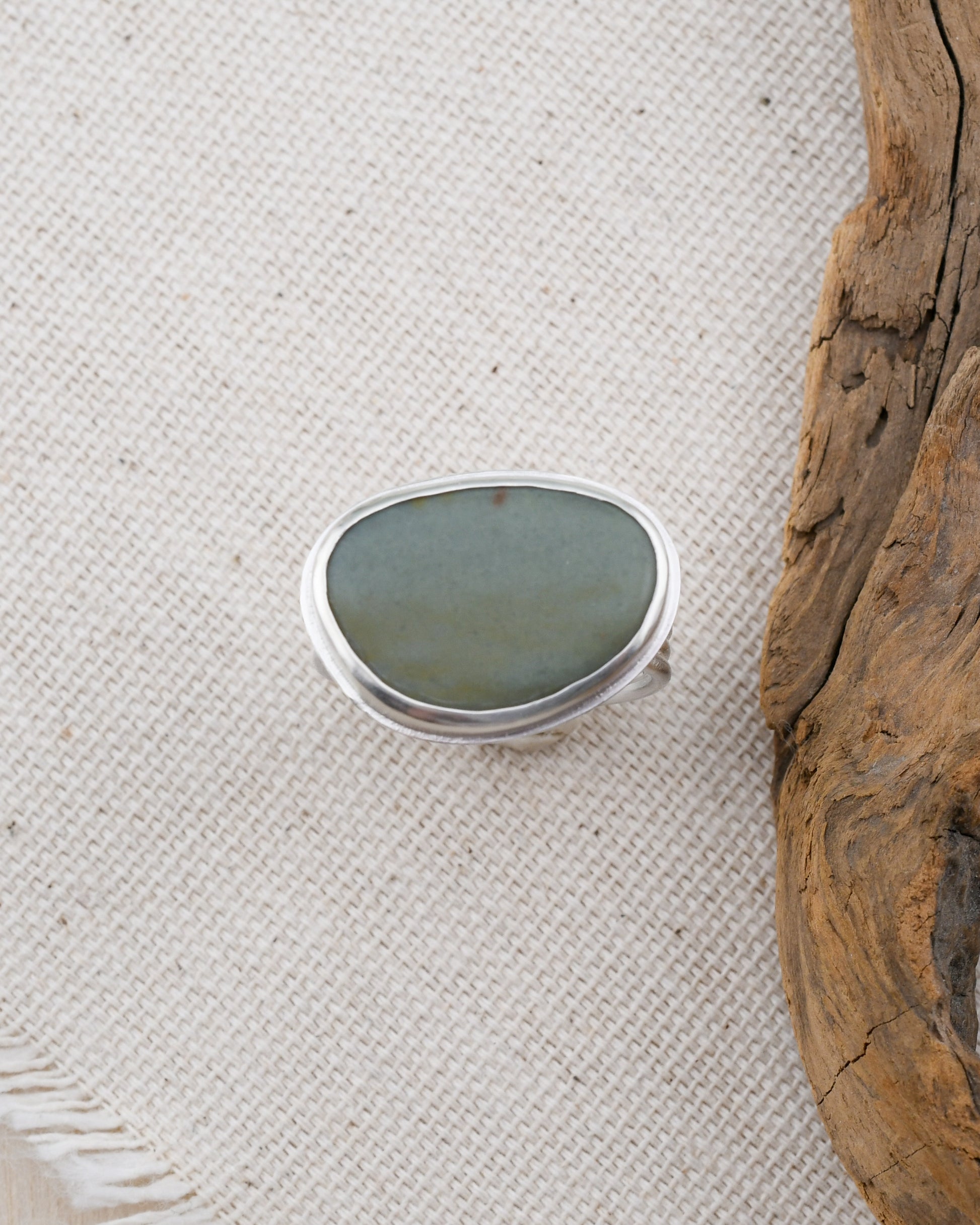 Silver ring with a gray-blue stone on a textured white surface with driftwood.