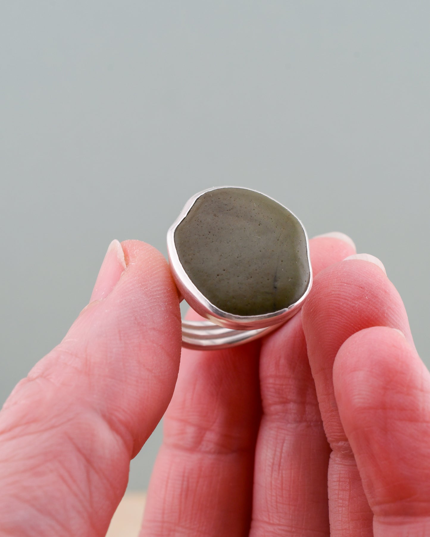 Hand holding a silver ring with a large green beach stone against a gray background