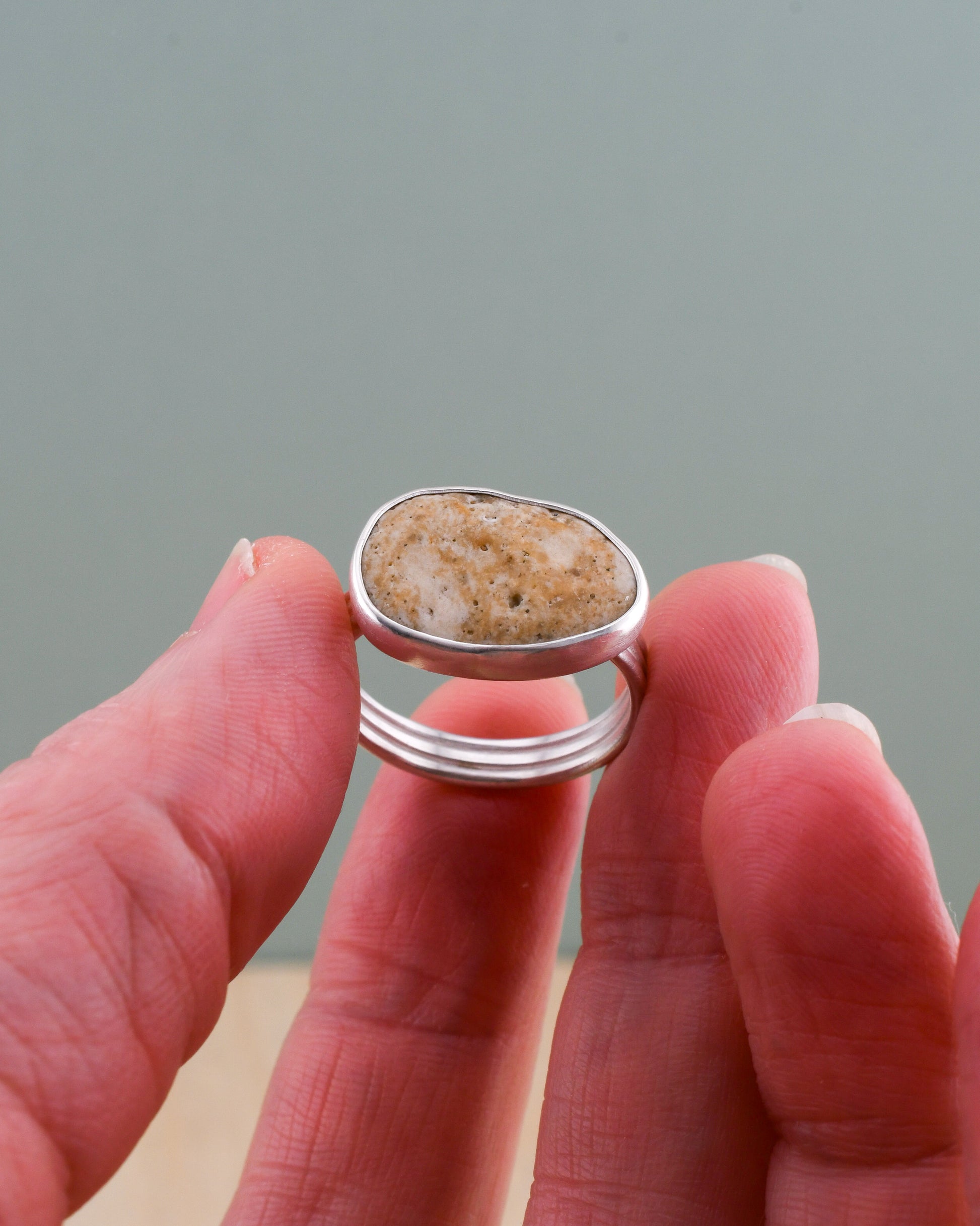 Hand holding a silver ring with a natural stone design against a gray background
