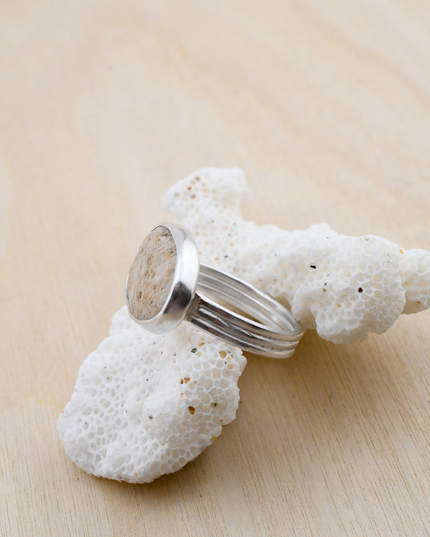 Silver ring with a textured beige beach stone displayed on white coral and pale wood background