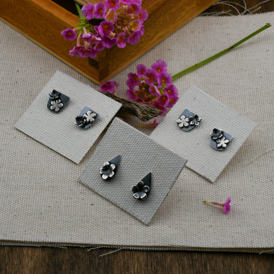 Three pairs of silver floral stud earrings displayed on small white cards over a beige fabric surface, with pink flowers and a wooden tray.