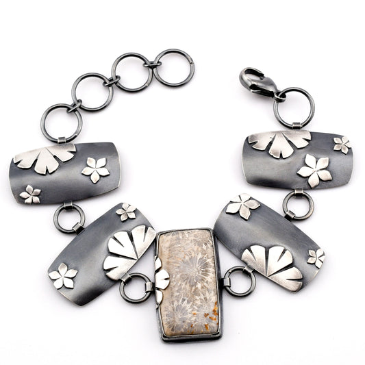 A linked bracelet with rectangular silver plates adorned with silver flower and a rectangle center stone of fossilized coral arranged in a circle on a white surface