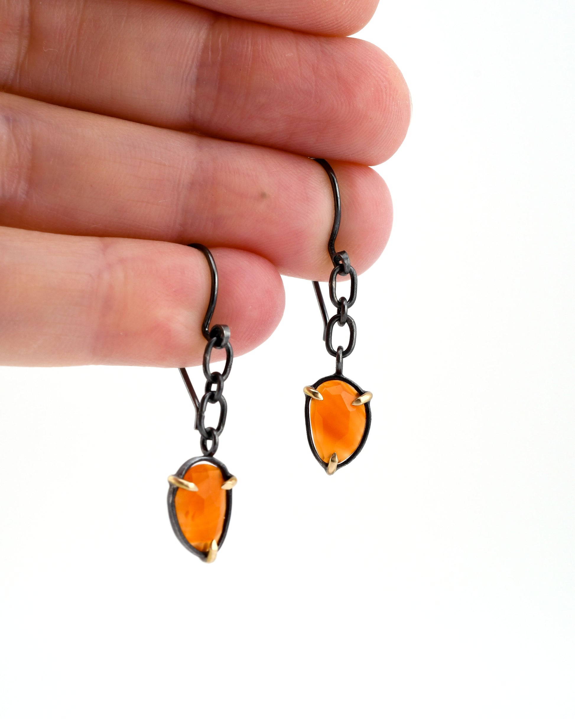 Orange teardrop earrings with black chain and gold prongs on a hand against a white background