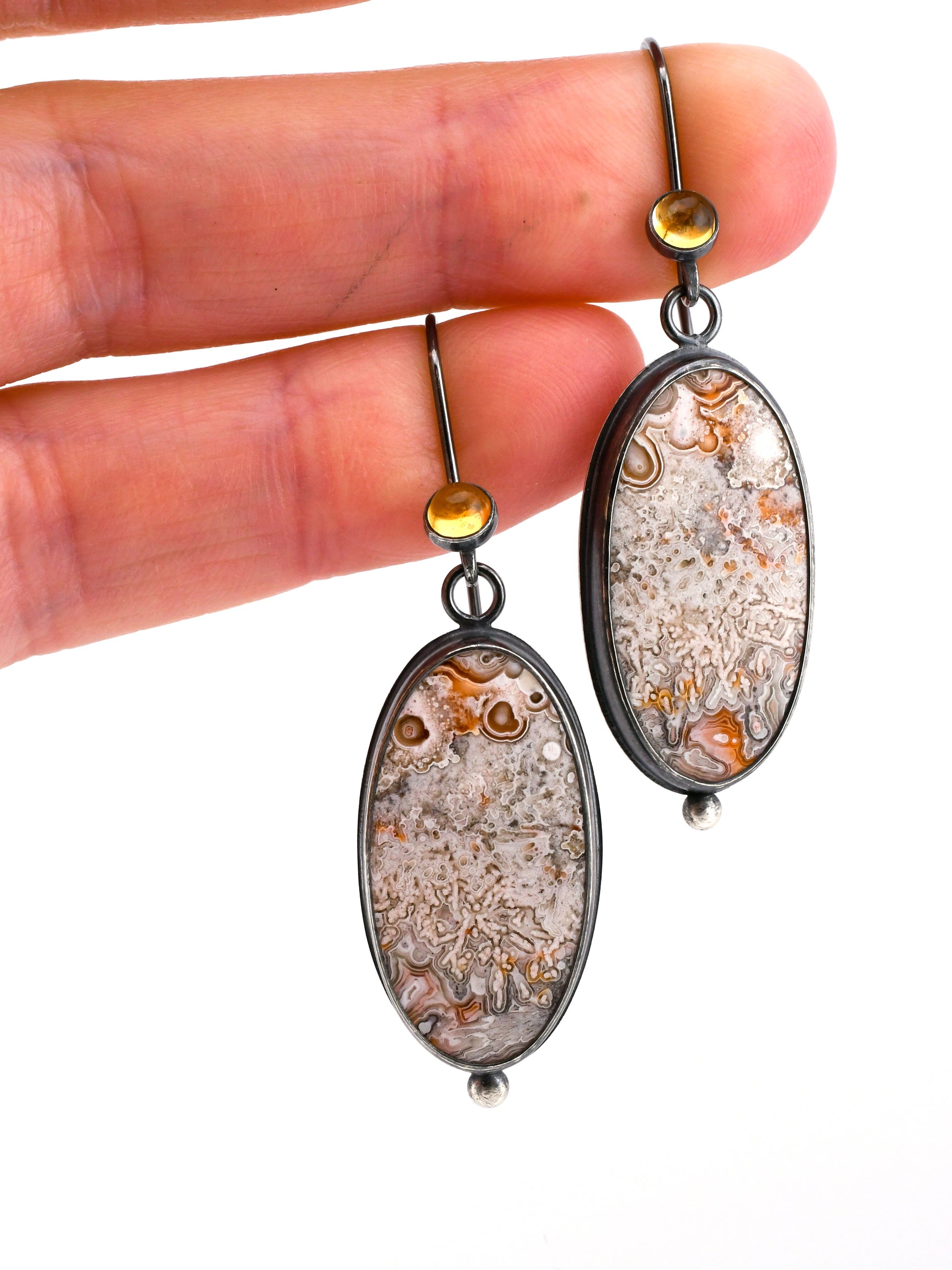 Oval-shaped earrings with intricate designs held between fingers against a white background