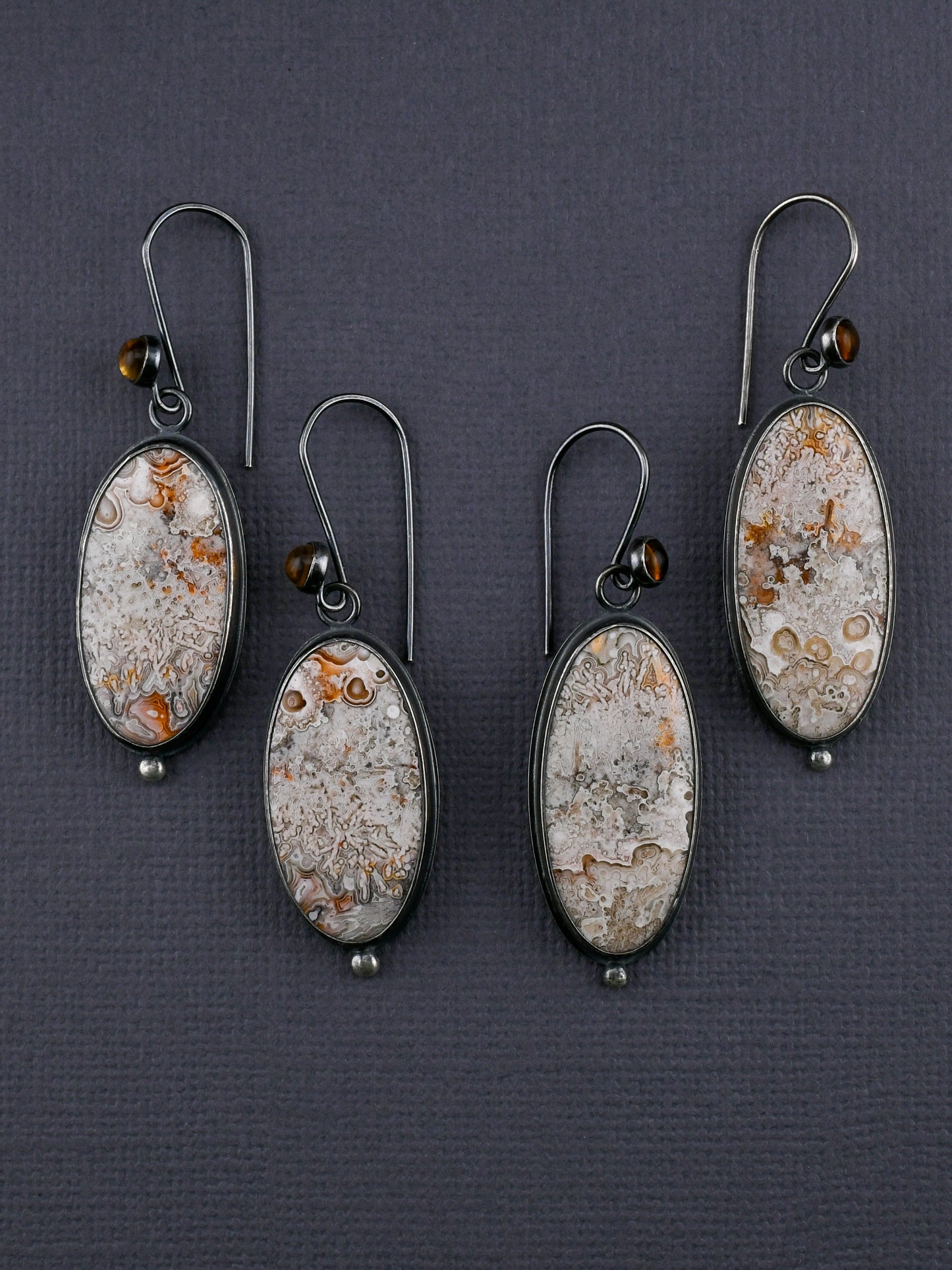 Two pairs of oval earrings with intricate designs on a dark gray background