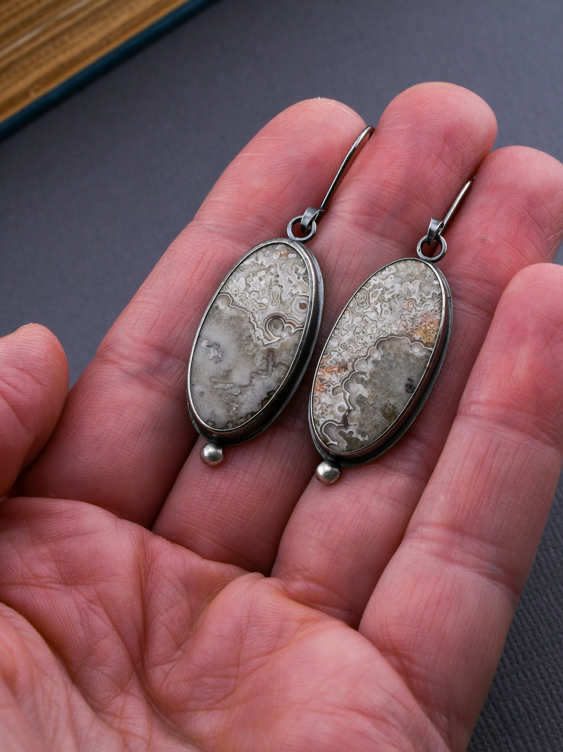 Two oval-shaped earrings with textured stones held in a hand against a dark background.