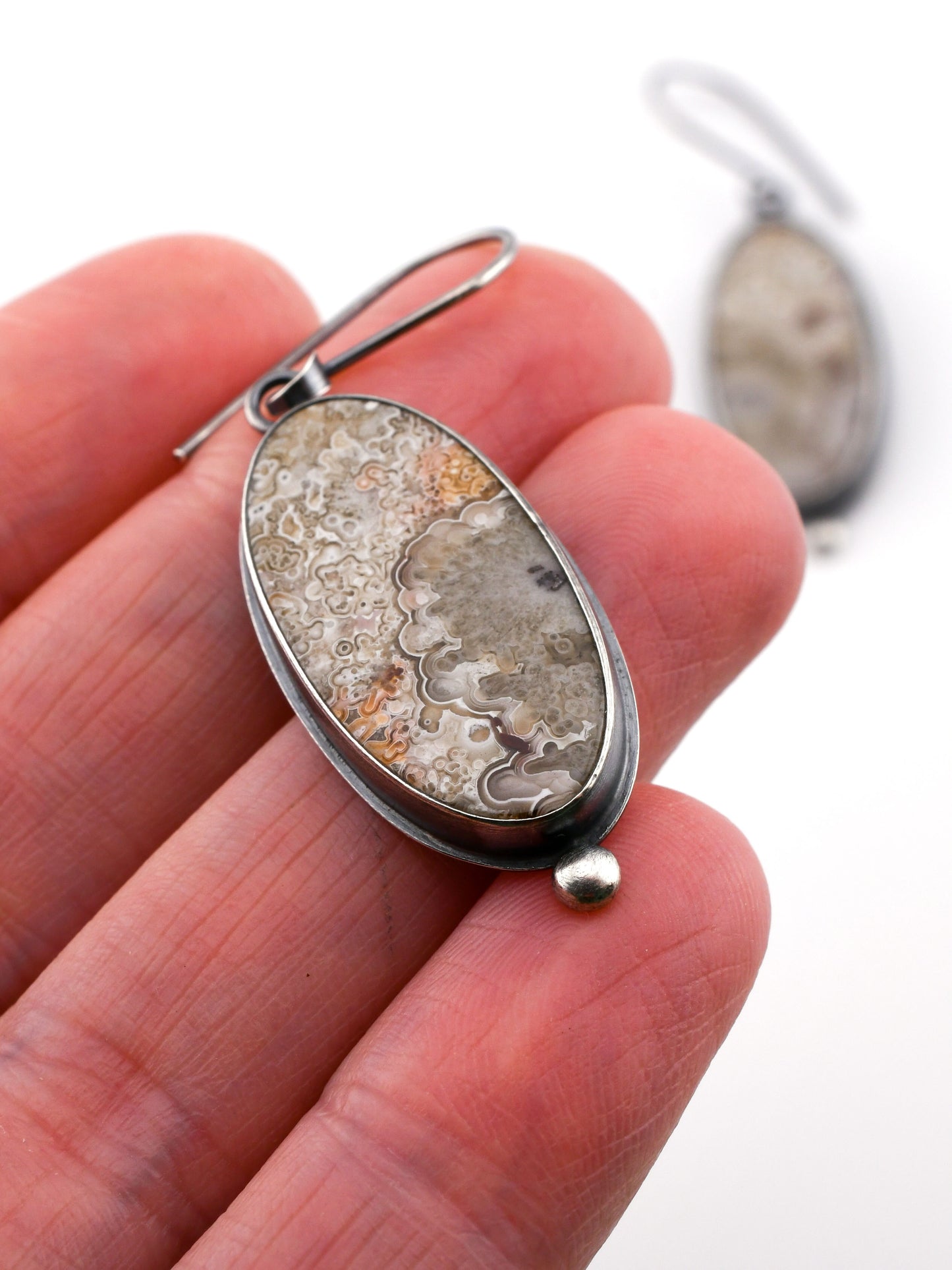 Oval-shaped silver earring with a natural stone held between fingers against a white background