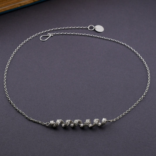 A woven silver coil pendant with silver chain displayed on textured gray surface with a book in the background