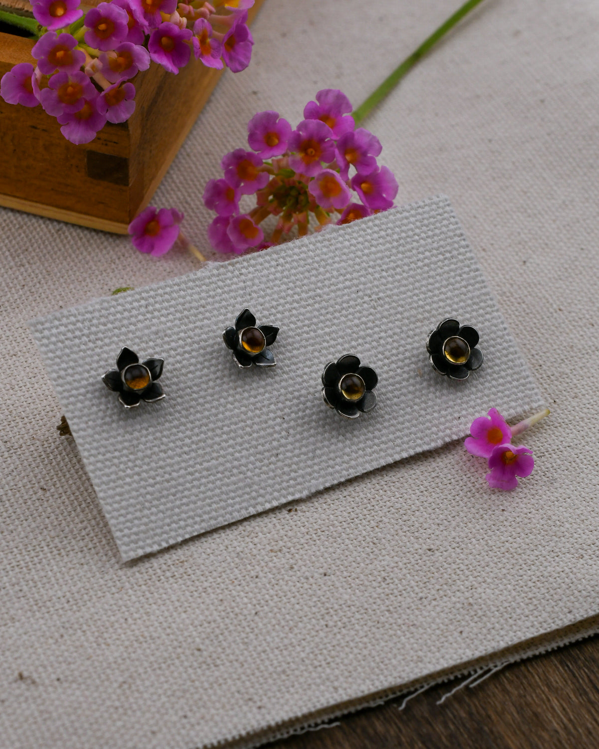 Two pairs of black flower-shaped stud earrings with yellow citrine centers, displayed on a textured white card over a beige fabric surface with small pink flowers in the background.