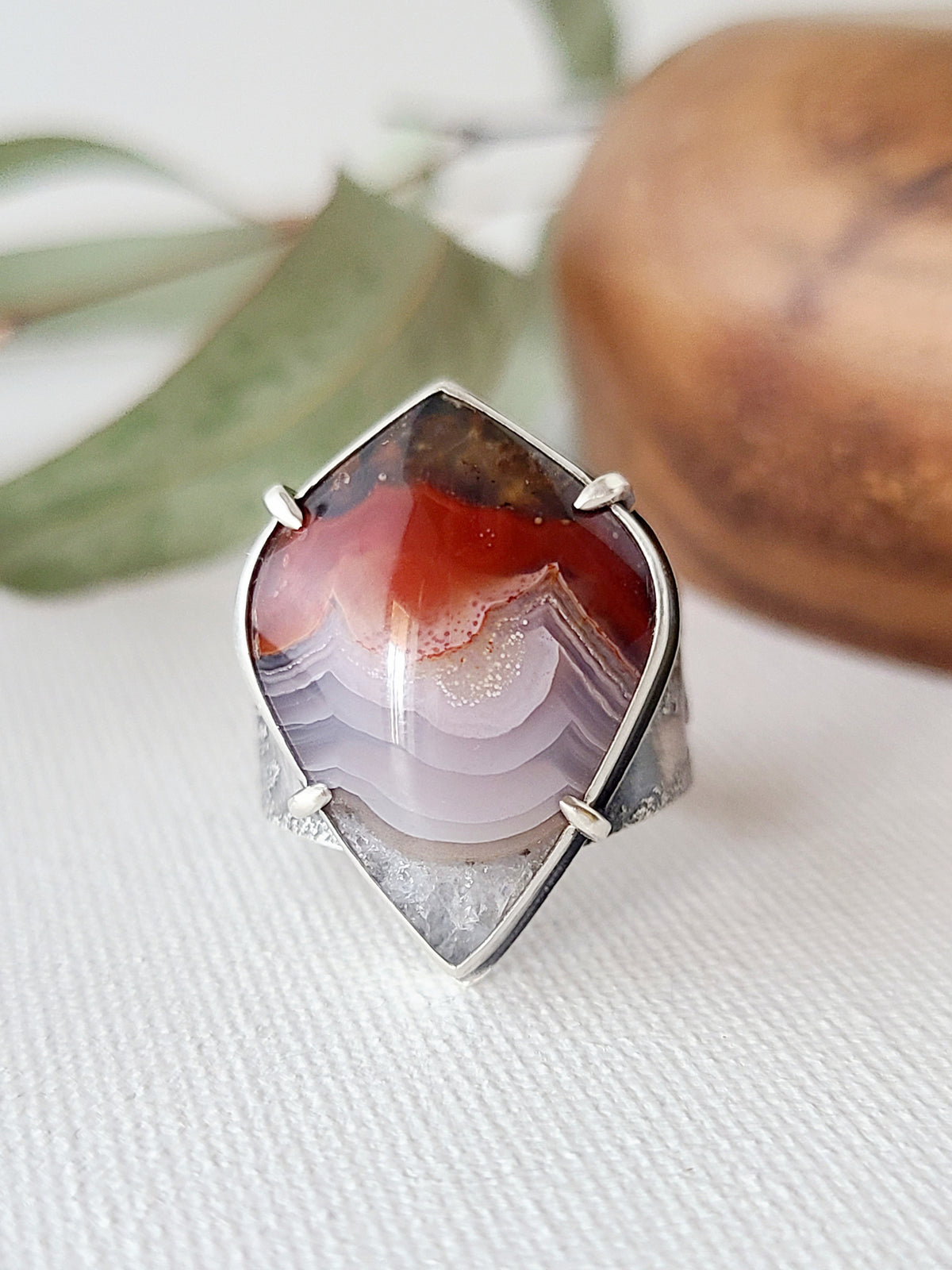 one of a kind statement ring with a prong set purple and red laguna lace agate shown on a textured white surface with leaves and dark wood in the background