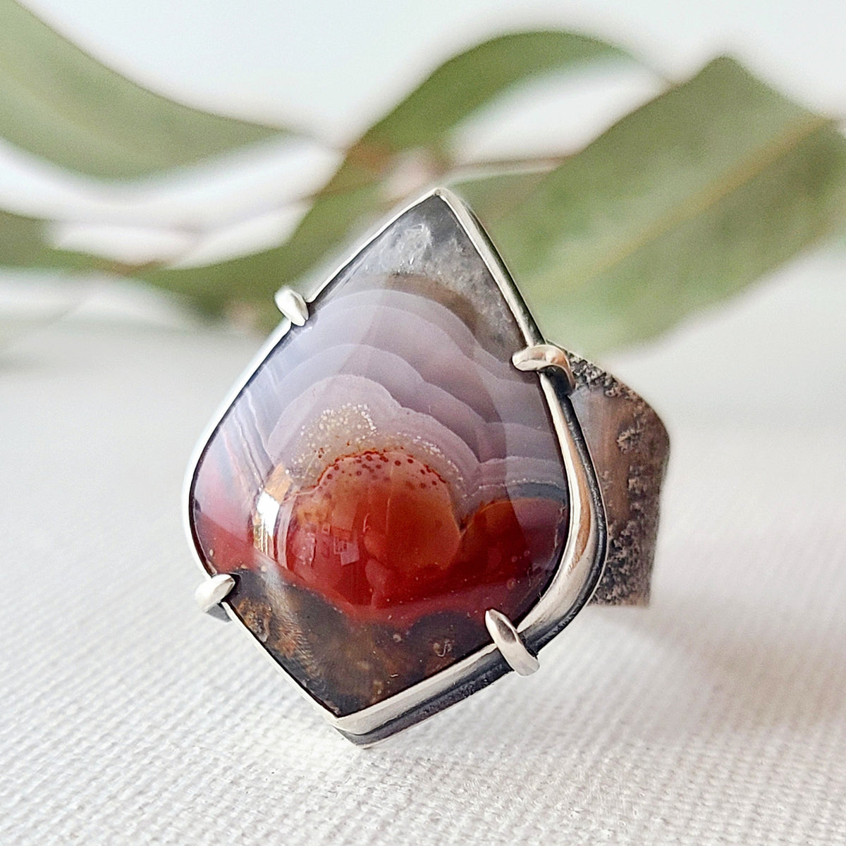 statement ring with a laguna lace agate in red and purple patterns set in a silver bezel with silver prongs on a textured band show on a textured white surface with green leaves in the background