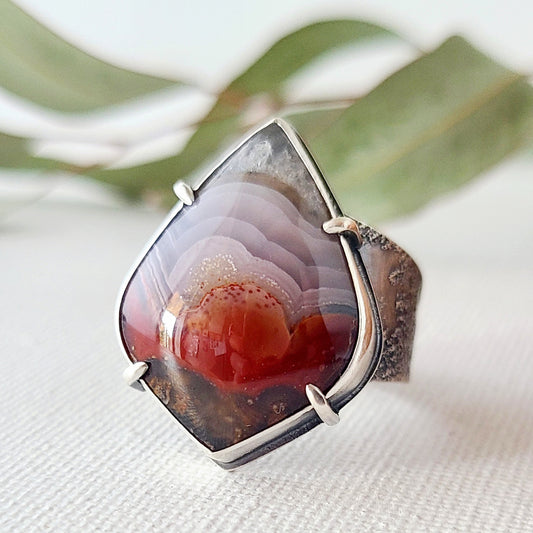 statement ring with a laguna lace agate in red and purple patterns set in a silver bezel with silver prongs on a textured band show on a textured white surface with green leaves in the background