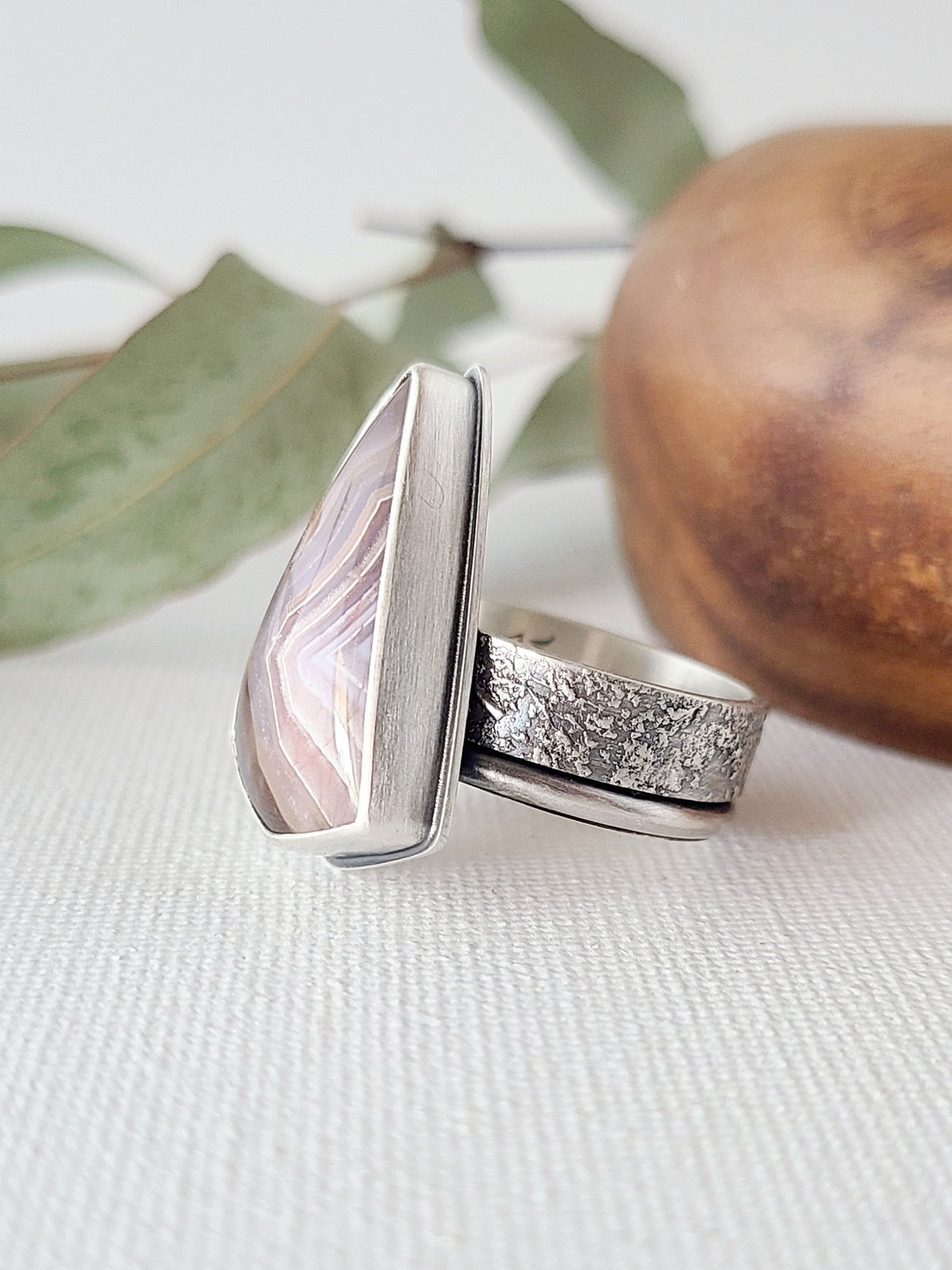 Side view of a large purple Laguna lace agate ring with a textured silver band shown on a white linen surface with leaves and a wooden bowl in the background