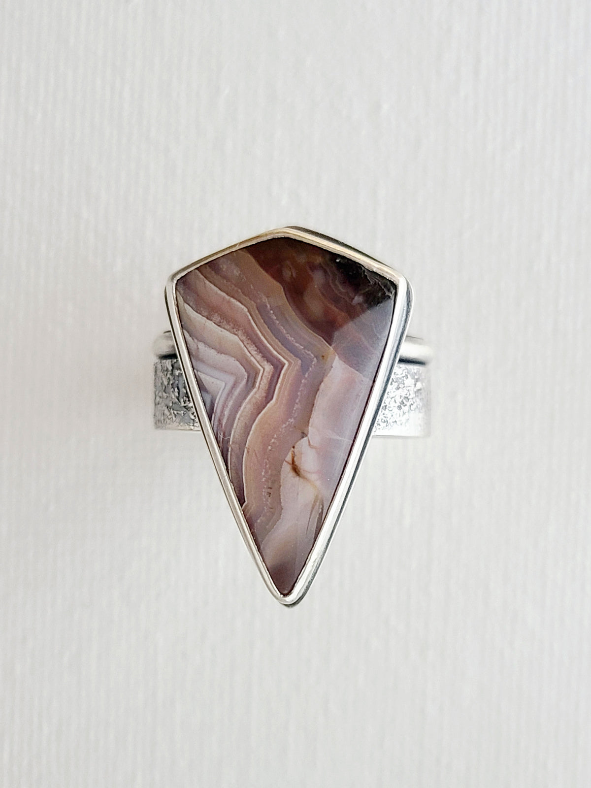 Top view of a unique statement ring featuring a large triangular Laguna lace agate with purple patterns and a textured silver back on a textured white surface