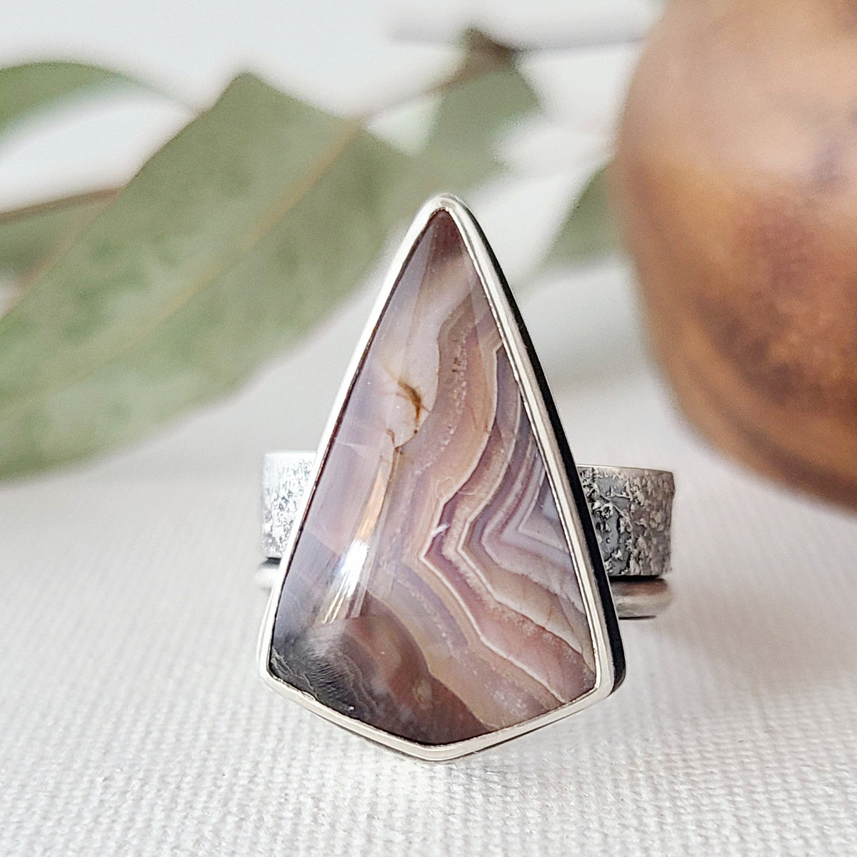 Triangular purple laguna lace agate statement ring with a textured silver band shown on a textured white surface with leaves and a wooden bowl