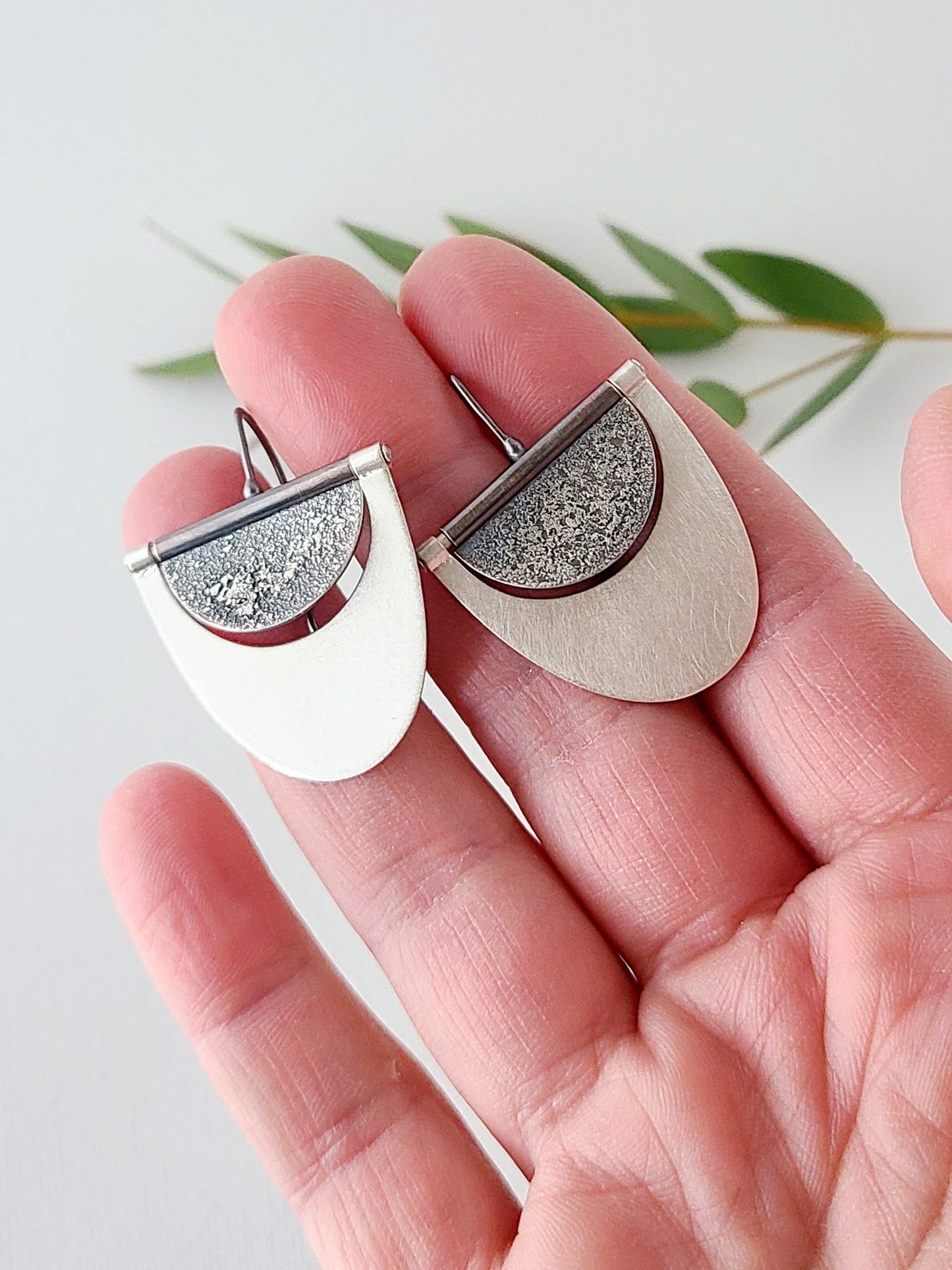 Silver earrings with half oxidized half bright silver surface held in a hand over a white surface and green leaves