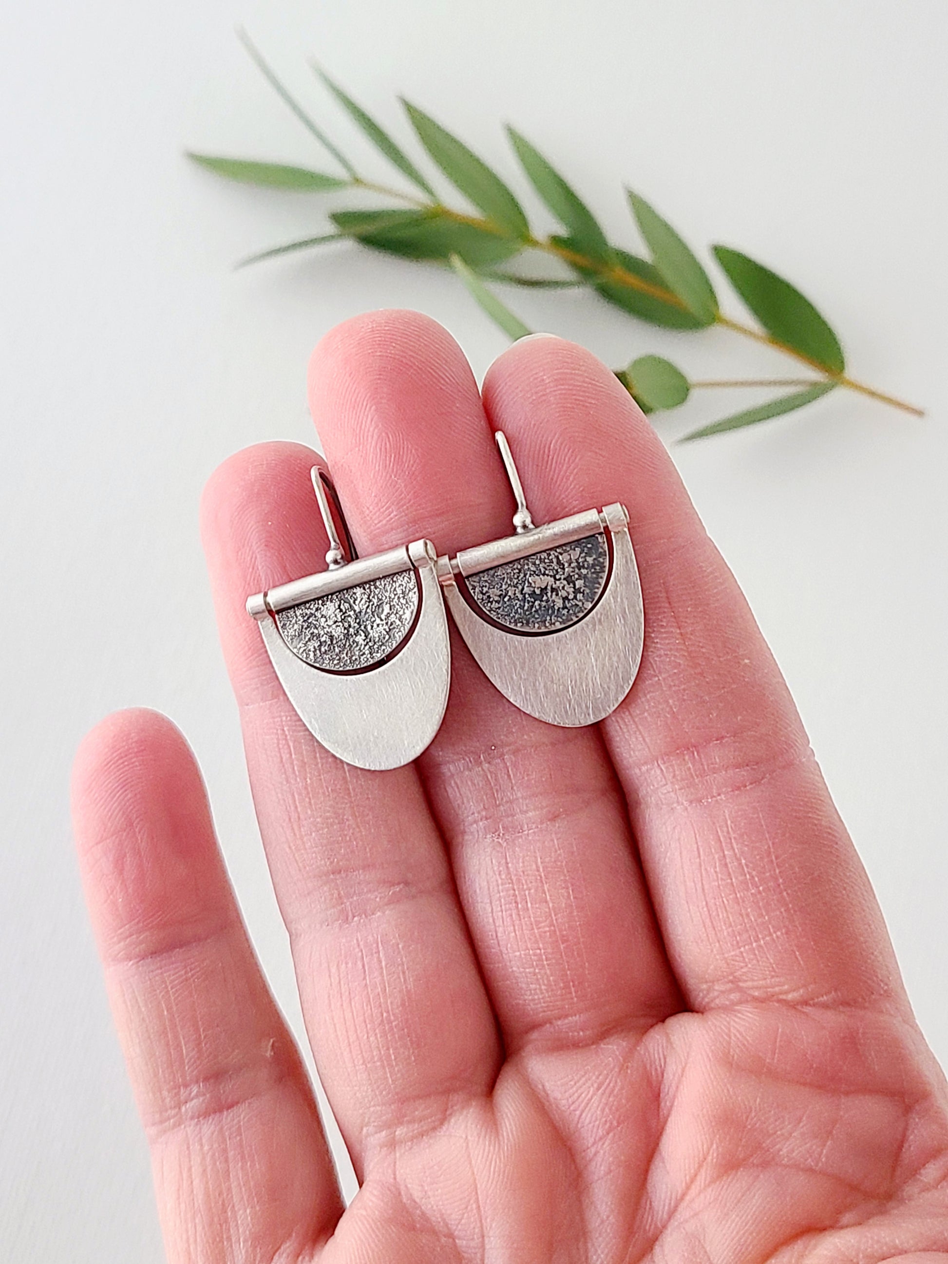 Silver earrings with half oxidized half bright silver surface held in a hand over a white background with green leaves