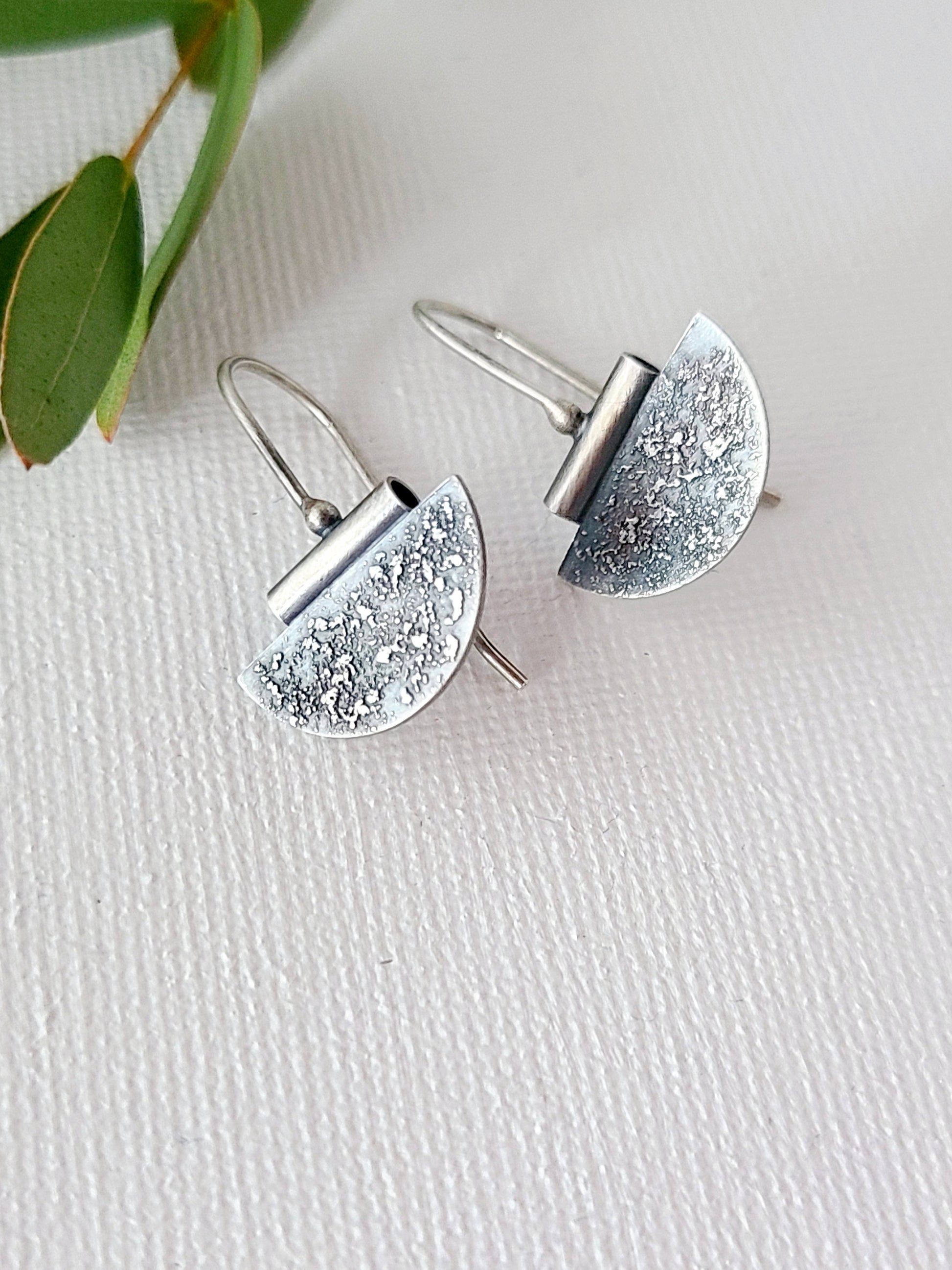 Textured and oxidized half-circle silver earrings on a textured white background with green leaves