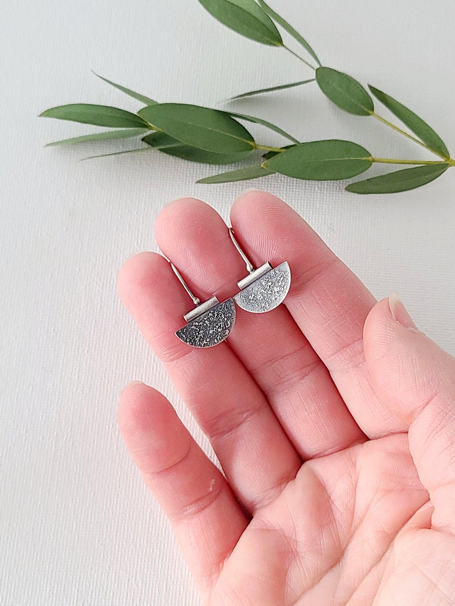 Small silver earrings in a half-circle shape with a textured and oxidized surface held in a hand over a white background and green leaves