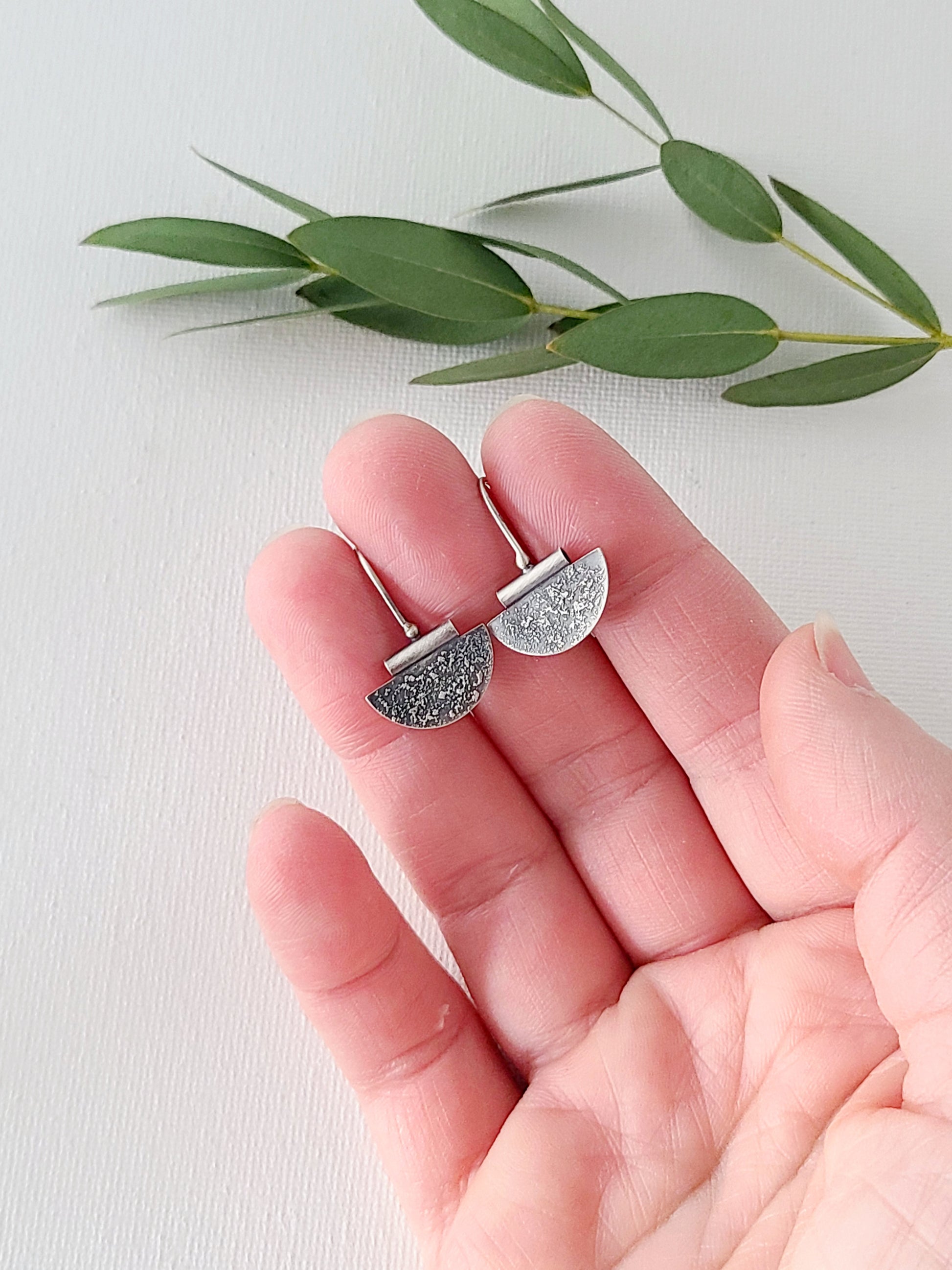 Small silver earrings in a half-circle shape with a textured and oxidized surface held in a hand over a white background and green leaves