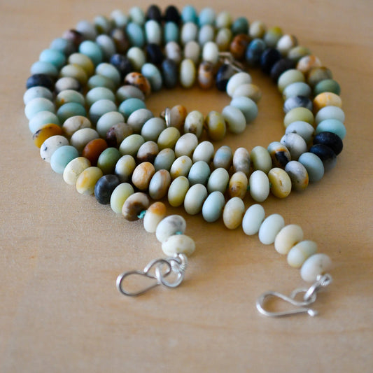 Two strands of knotted beaded necklaces with matte-finished Amazonite beads in various shades, including blue, beige, and green, displayed on a wooden surface. Each necklace ends with a silver clasp.