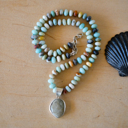 A hand-knotted Amazonite and Beach Stone Necklace with a silver pendant featuring a light blue streaked stone, displayed on a wooden surface alongside a black shell.