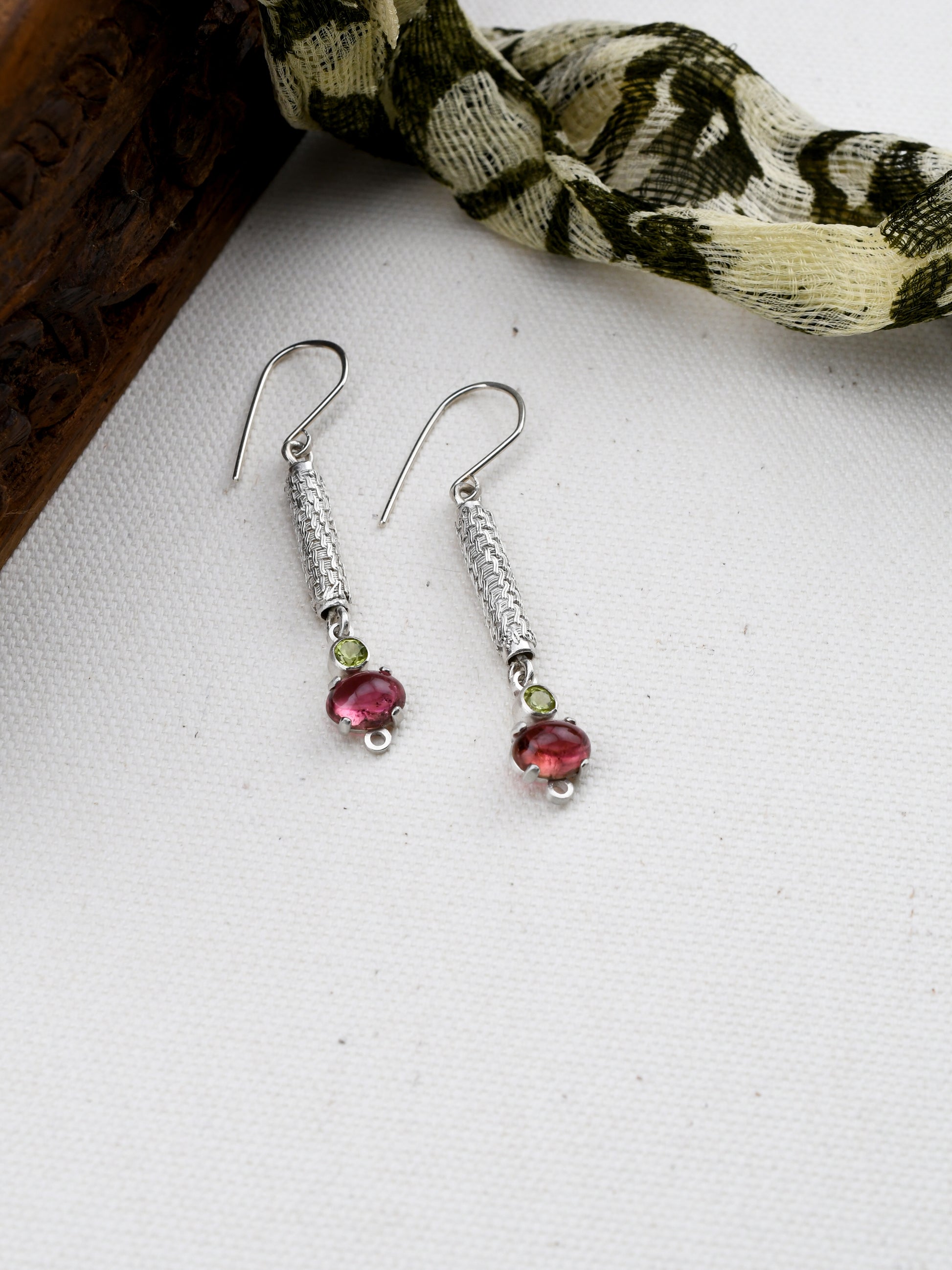 Silver earrings with pink and green stones on a white surface with a patterned fabric background.