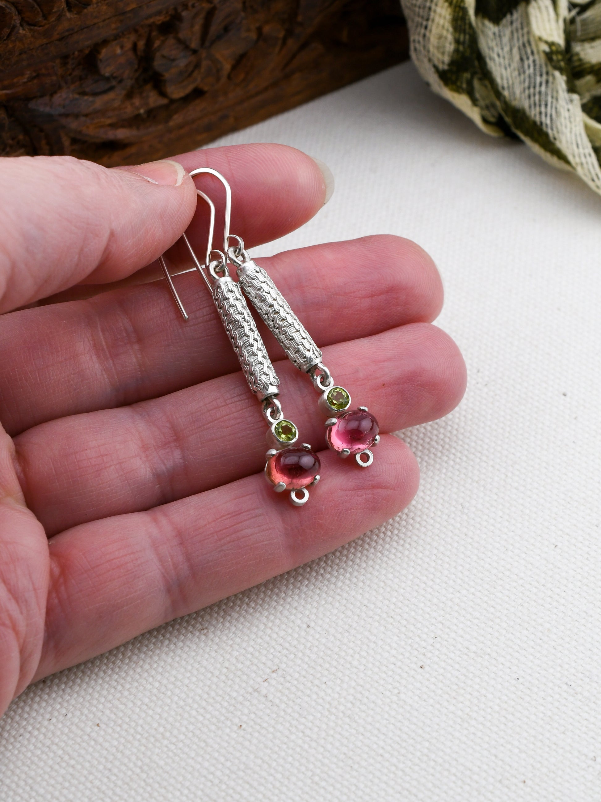 Silver earrings with pink and green gemstones held between fingers on a white surface.