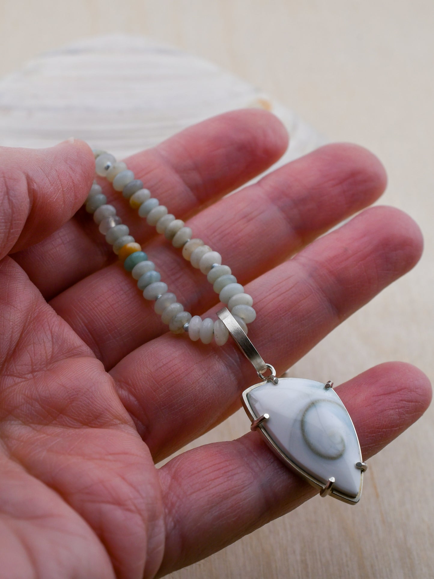 Hand holding a beaded necklace with a spiral shell pendant on a light background