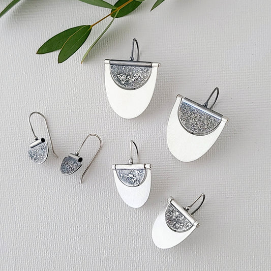 A collection of silver earrings in three different sizes displayed on a grey surface, with the largest pair at the top and the smallest at the bottom, resembling a dangle design with a textured finish.