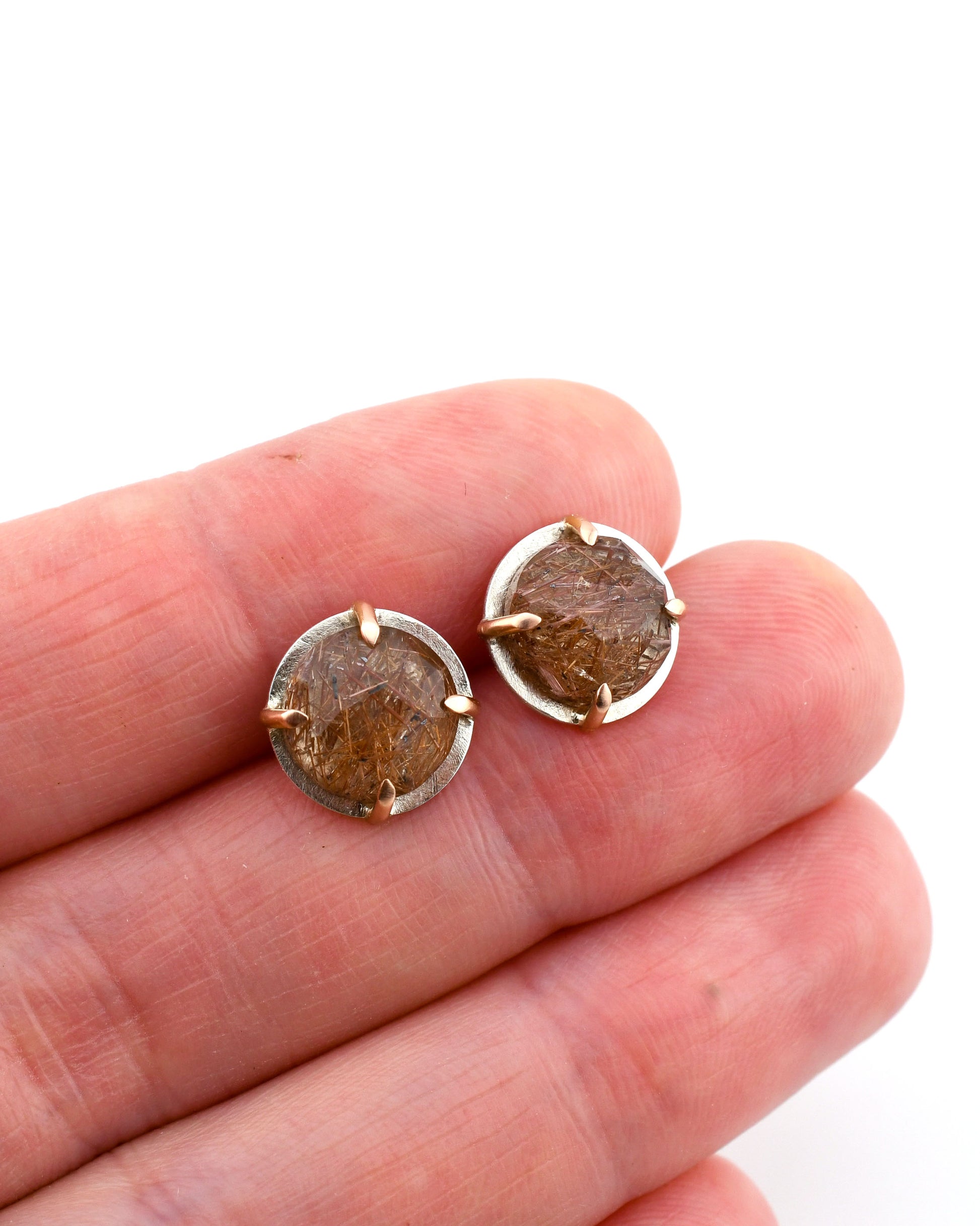 silver and rose gold round stud earrings with red rutilated quartz held between fingers over a white background
