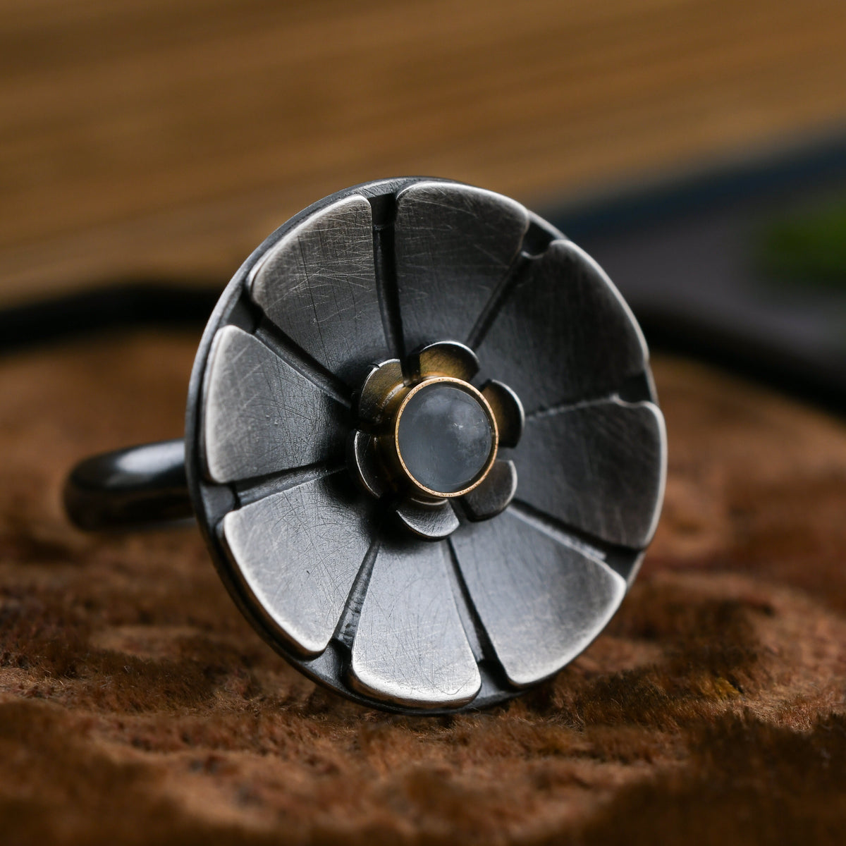 A close-up of a handmade flower ring made of oxidized silver with a central round pale blue aquamarine stone and a small gold accent, resting on a textured brown surface.