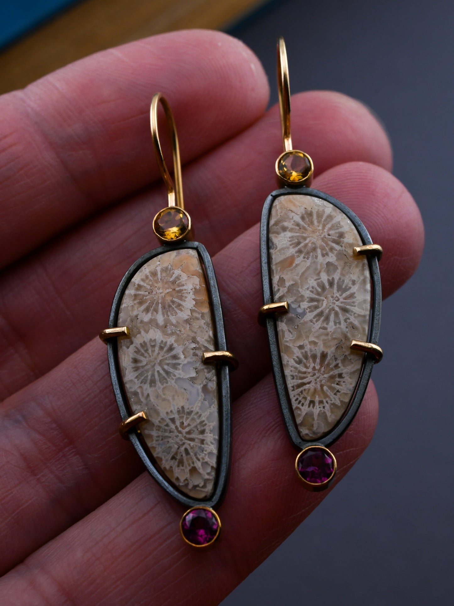 Fossilized coral earrings with gold and gemstone accents held in a hand against a dark background.