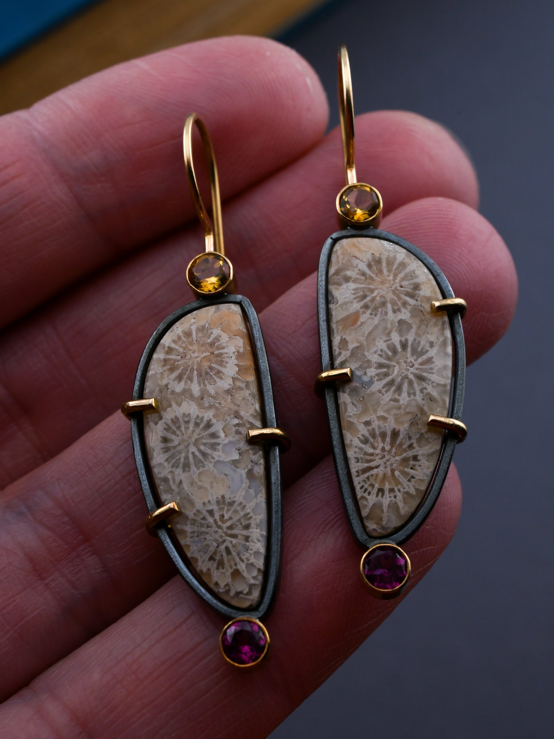 Fossilized coral earrings with gold and gemstone accents held in a hand against a dark background.