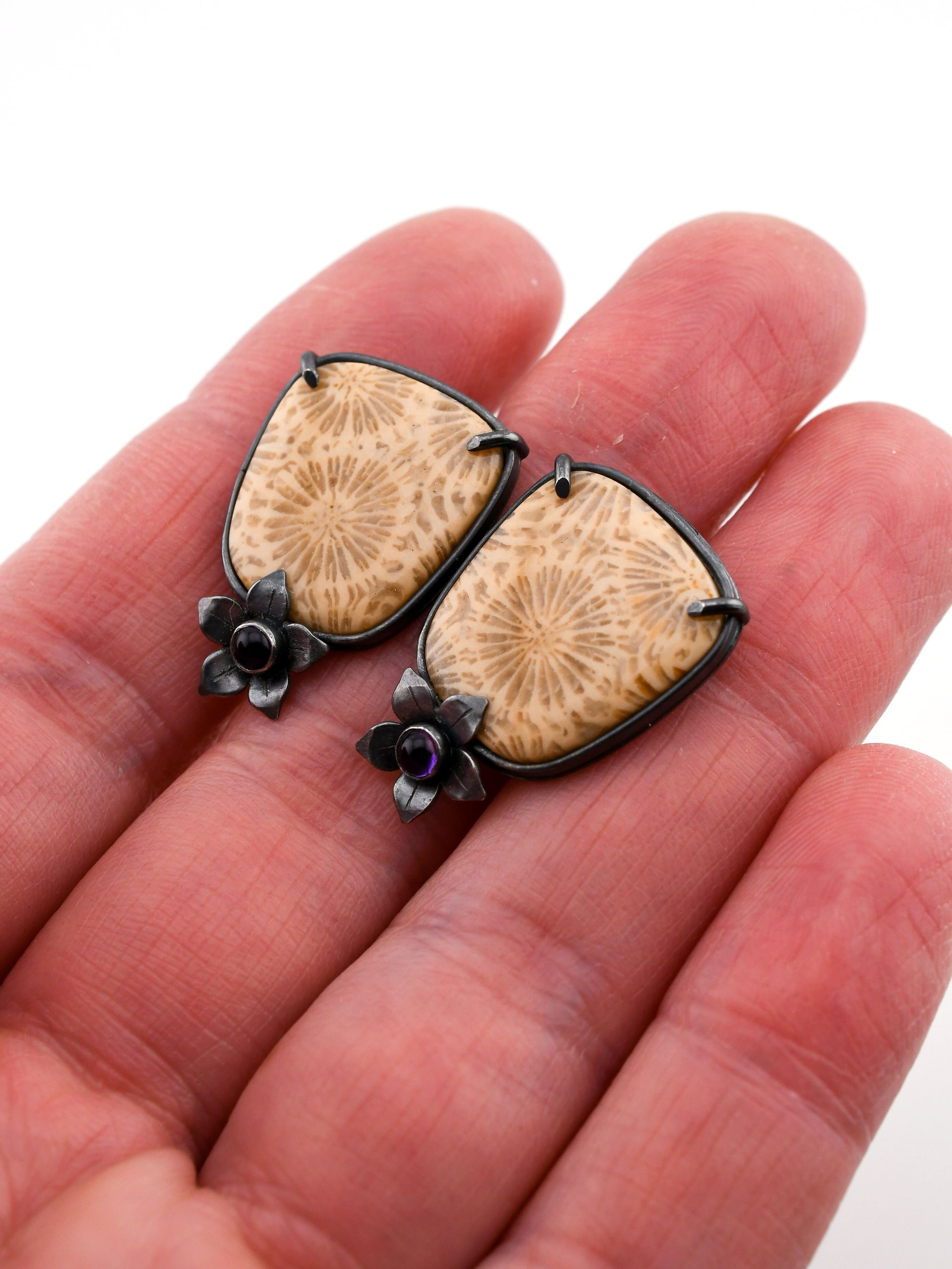 Fossilized coral earrings with black floral accents held between fingers on a white background