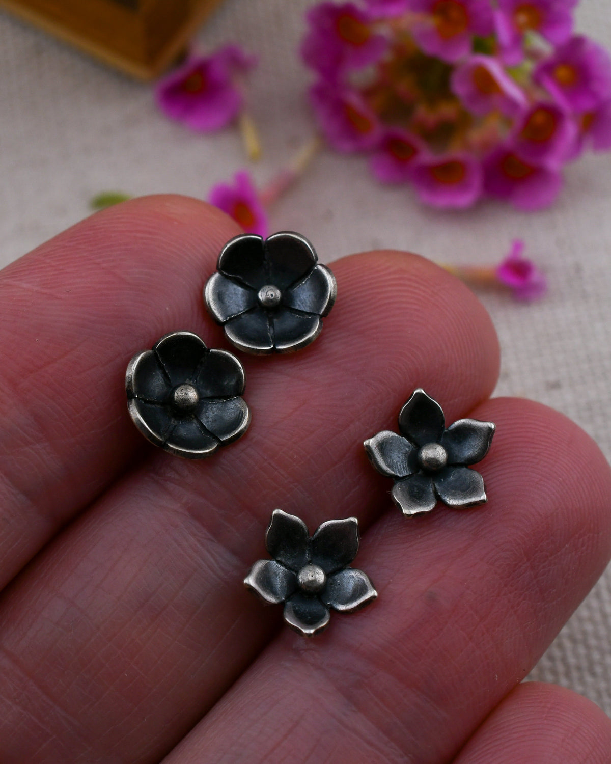 Two pairs of small flower-shaped stud earrings, held in person's fingers against a soft beige fabric background with blurred pink flowers.