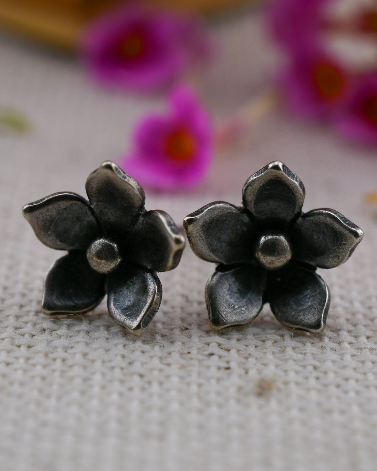 A pair of dark oxidized silver jasmine flower stud earrings with five petals, resting on a textured beige fabric with blurred pink flowers in the background.