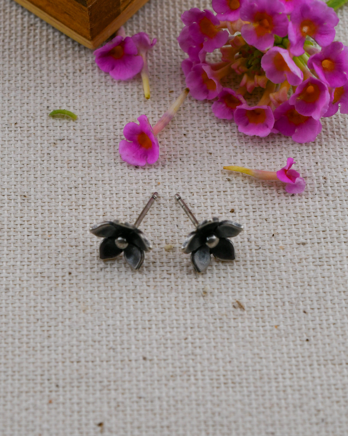 A pair of small blackened silver jasmine flower-shaped stud earrings, displayed on a textured beige fabric background with pink flowers.