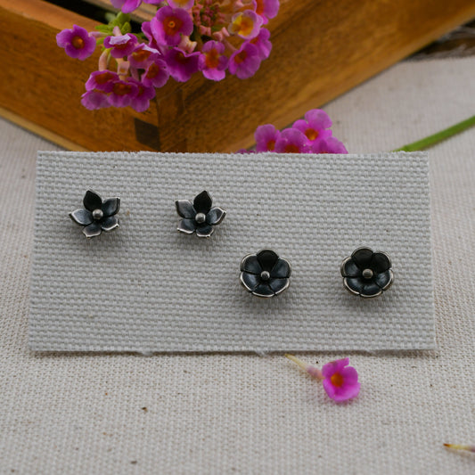 Two pairs of small black flower-shaped stud earrings with silver button centers, displayed on a rectangular textured gray card. The card rests on a light fabric surface with pink flowers and a wooden box in the background.