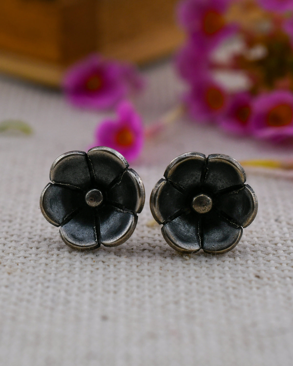 A pair of silver poppy flower-shaped stud earrings resting on a textured beige fabric surface with blurred pink flowers in the background.