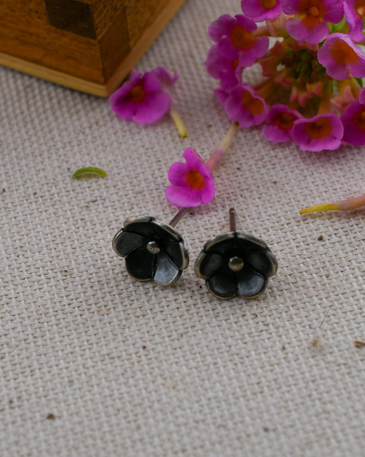 A pair of blackened silver poppy flower-shaped stud earrings, displayed on a textured beige fabric surface with pink flowers and a wooden box in the background.