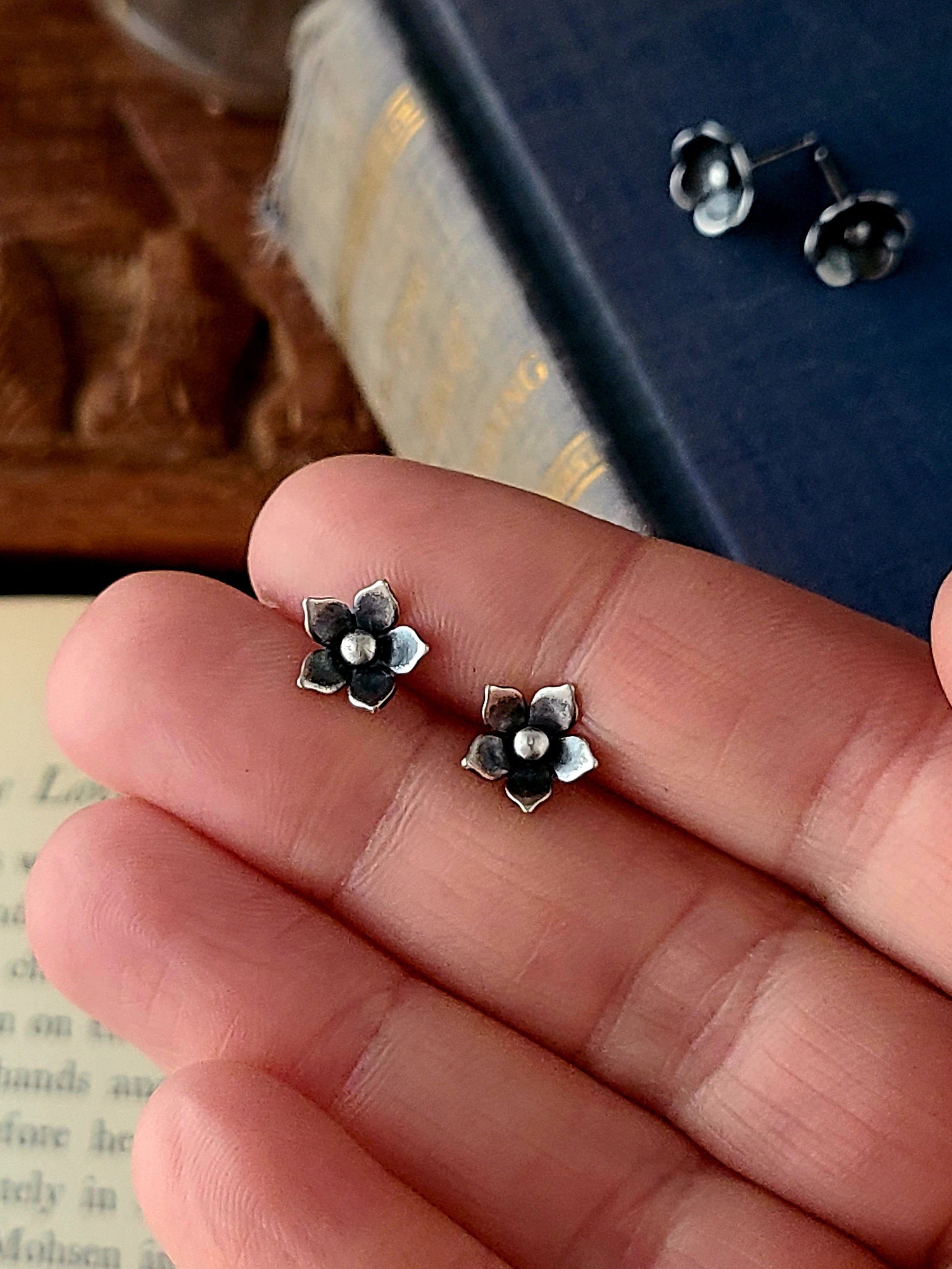 silver flower stud with five petals held in a hand over a book and a wood box