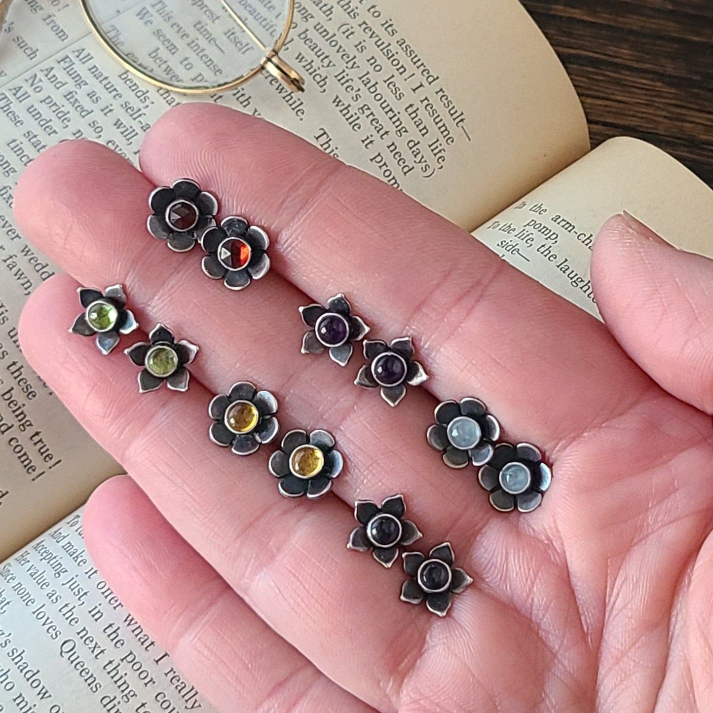 Hand holding six pairs of flower studs with colorful gemstone centers over an open book with a dark wood background