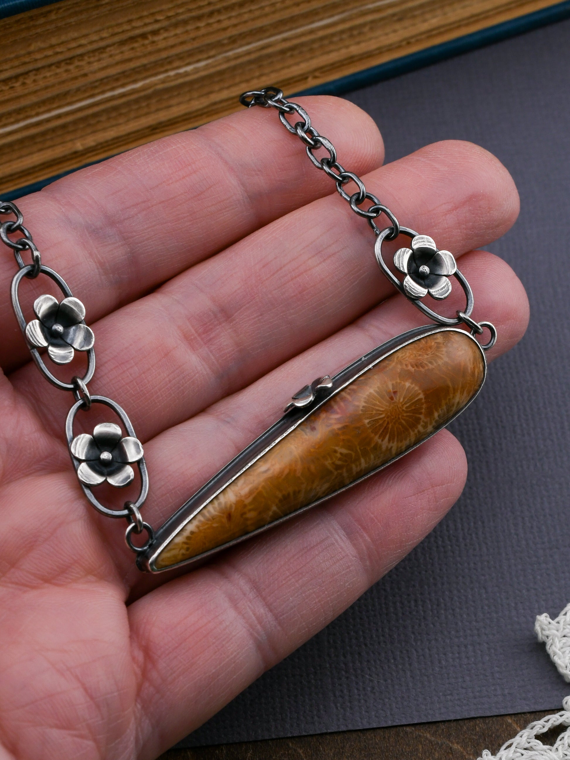 Hand holding  long golden fossil coral pendant with blackened silver chain and silver flower elements on a gray background