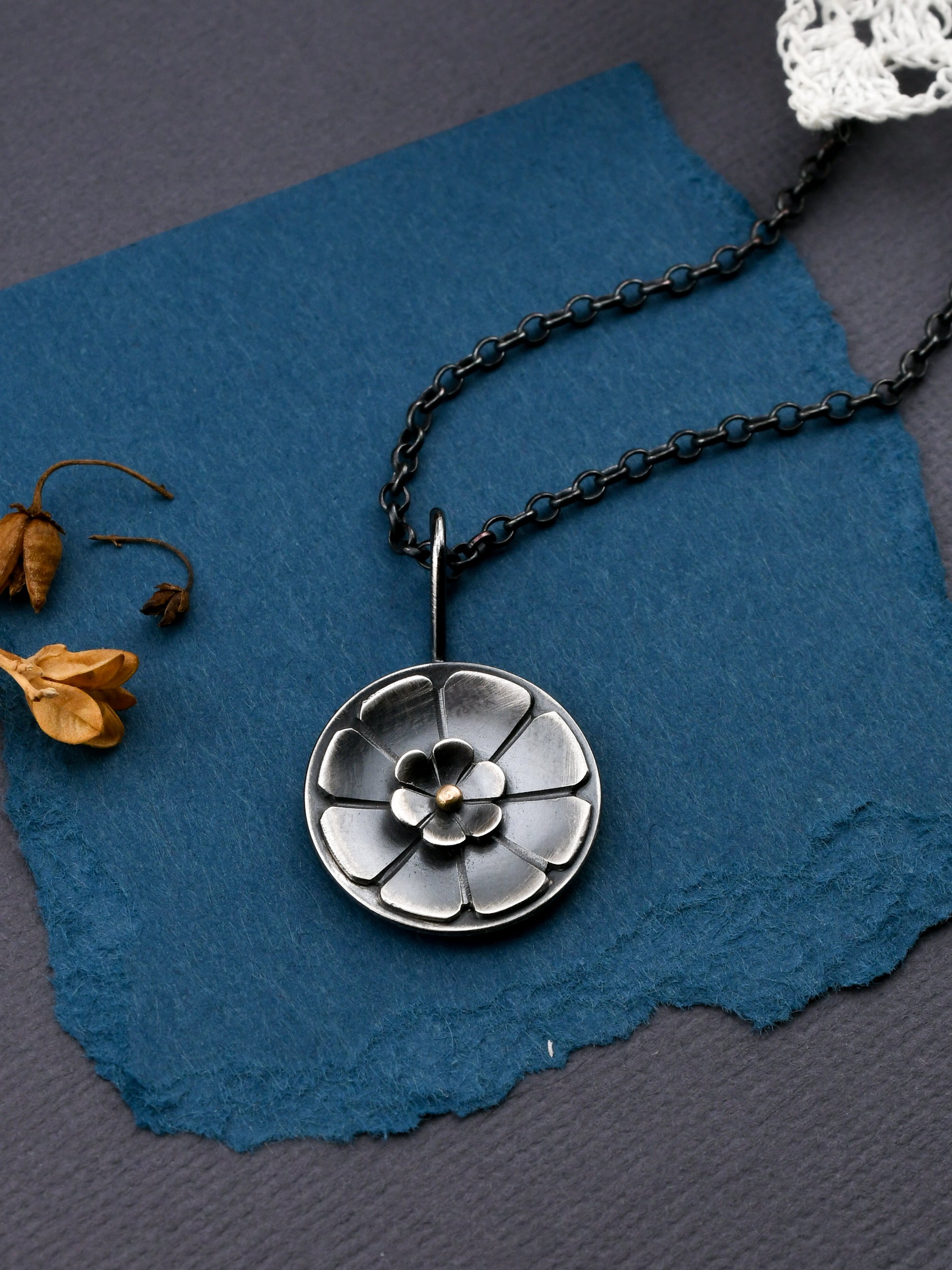 Silver floral pendant necklace on a blue textured surface with dried flowers.