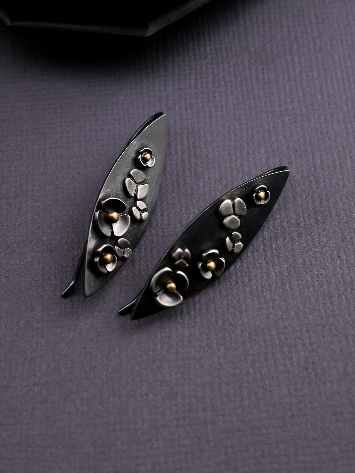 Leaf shaped earrings of blackened silver with floral details and gold accents on a dark textured background